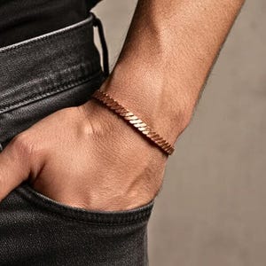 Peut inclure: Un bracelet de couleur cuivre avec un motif de maillons de chaîne. Le bracelet est porté au poignet, et la personne porte un jean noir. Le bracelet mesure environ 1,3 cm de large.