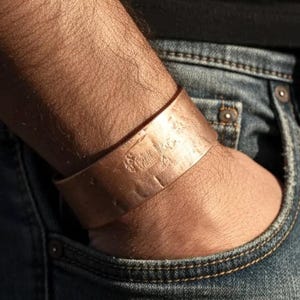 May include: A wide copper cuff bracelet with a textured surface. The bracelet is worn on a wrist, with the arm resting in a blue denim jeans pocket. The cuff is a warm, metallic colour.
