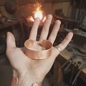 May include: A copper cuff bracelet and a matching wrist cuff. The bracelet is held in a hand, with a second cuff on the wrist. The background shows a blacksmith's workshop with a forge and tools.