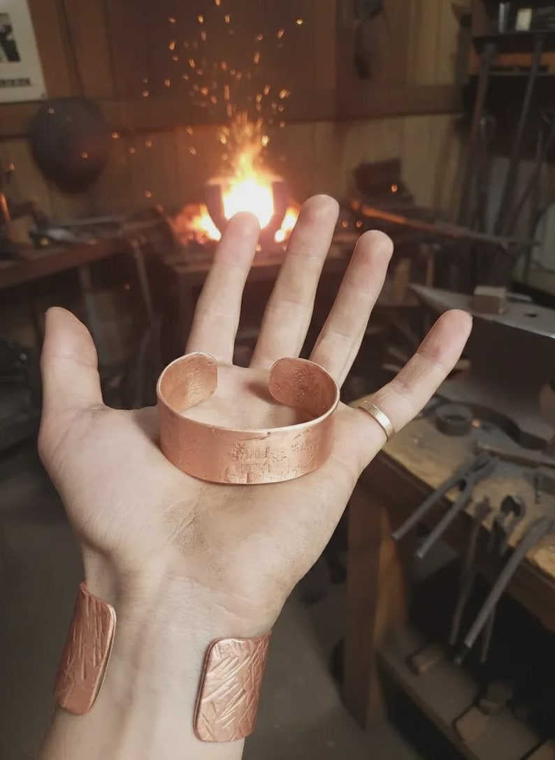 May include: A copper cuff bracelet and a matching wrist cuff. The bracelet is held in a hand, with a second cuff on the wrist. The background shows a blacksmith's workshop with a forge and tools.