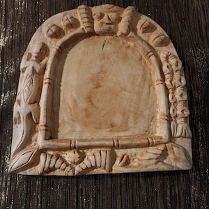 May include: A light beige, arched wall plaque featuring a central recessed area framed by carved details. The border includes a lizard figure on the left, a vertical row of circular shapes on the right, and a stylised face at the top centre.