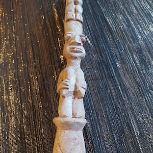 Puede incluir: Una talla de madera de color claro que presenta una figura de pie con una cabeza y rasgos faciales prominentes, rematada por una aguja alta, puntiaguda y segmentada. La escultura tiene una textura tallada y descansa sobre una base cilíndrica.