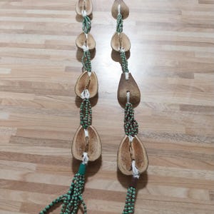Pode incluir: Um colar artesanal com vagens de sementes naturais e pequenas contas verdes. O colar tem um design longo e fluido com vários fios de contas e vagens de sementes. Pequenas conchas brancas são presas na parte inferior do colar.