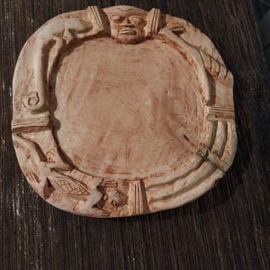 Puede incluir: Un plato circular de arcilla de color claro con un borde en relieve tallado a mano. El borde muestra figuras estilizadas, incluyendo un rostro central en la parte superior, formas animales alargadas y patrones geométricos.