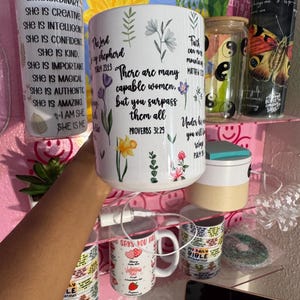 May include: A white ceramic mug with floral illustrations and the text "There are many capable women, but you surpass them all" from Proverbs 31:29. The mug is surrounded by other decorative items on a pink shelf.