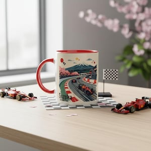 Puede incluir: Taza de cerámica blanca con interior y asa rojos. La taza presenta una colorida ilustración de un circuito de carreras con coches, el monte Fuji y flores de cerezo. También es visible el texto "Suzuka Grand Prix".