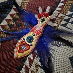 May include: A decorative object featuring a central painted earth globe, a red heart with an eye, and three clear quartz points. It is adorned with small red roses, white pearls, and dark blue feathers extending from the sides and top.