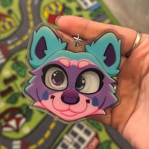 May include: A colorful cartoon animal face keychain. The keychain features a smiling face with blue ears, purple fur, and a pink nose and mouth. The keychain is held in a hand against a colorful play mat.