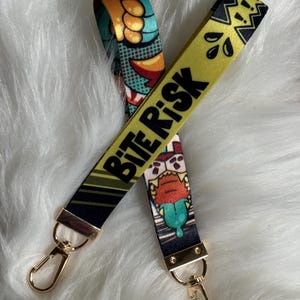 May include: A fabric lanyard featuring a yellow and black color scheme with the text BITE RISK printed in bold black letters. The design includes comic-style graphics of a creature with sharp teeth and a green tongue, finished with gold metal clasps.