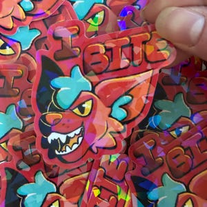 Könnte beinhalten: Eine Ansammlung roter, holografischer Aufkleber mit einem Cartoon-Fuchs mit grimmigem Gesichtsausdruck. Die Aufkleber tragen den Text "I BITE" mit blauen und gelben Akzenten. Eine Hand hält die Aufkleber.