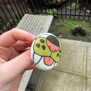 May include: A round, white button with a cartoon design featuring green, black, and pink elements. The button has a soft, fabric-like texture and is held in a hand.