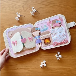 May include: A felt quiet book with a bear and doll theme. The open book reveals a bear face, a doll, a bed, and a fireplace. The book is in shades of pink, white, and purple, with floral accents and a button closure. Small white flowers are scattered around.