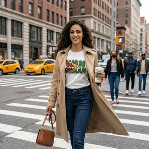 May include: A woman wearing a white t-shirt with the text "WAH GWAAN" in red, yellow, and green, paired with blue jeans and a tan trench coat. She carries a brown purse and a coffee cup. The background shows a city street with yellow taxis.