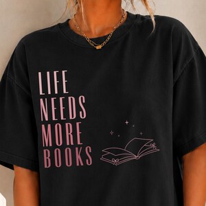 May include: A black short-sleeved t-shirt featuring the light pink text "LIFE NEEDS MORE BOOKS" printed vertically on the left side. Below the text is a simple line drawing of an open book with small star accents and ribbon details.