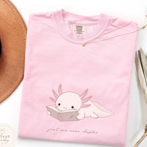 May include: A light pink crewneck t-shirt featuring a graphic of a pale pink axolotl reading an open book. Below the axolotl, the text reads "just one more chapter" in a cursive font. The shirt has a Comfort Colors brand label at the neck.