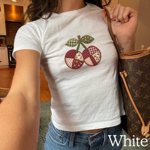 May include: A white short-sleeved t-shirt featuring a central graphic of two cherries designed with a patchwork pattern. The cherries are composed of various red and white fabric-style prints, while the stems and leaves are green with a checkered pattern.