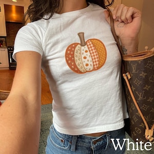 May include: A white short-sleeved t-shirt featuring a central graphic of a pumpkin composed of various vertical panels with different orange, cream, and brown patterns, including floral, gingham, and small pumpkin motifs, paired with blue denim jeans.