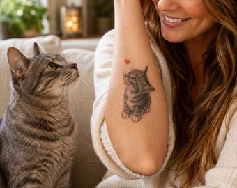 May include: A temporary tattoo on an arm featuring a detailed black and gray illustration of a small tabby kitten looking upward toward a tiny red heart symbol. The kitten has visible stripes and pointed ears, matching the appearance of the cat nearby.