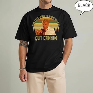 May include: Black t-shirt with a graphic of a man holding a drink, set against a sunset design. The text reads "LOOKS LIKE I PICKED THE WRONG WEEK TO QUIT DRINKING". A speech bubble in the upper right corner says "BLACK".
