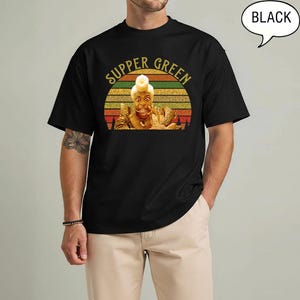 May include: Black t-shirt with the text "SUPPER GREEN" in a retro font above a graphic of a person. The graphic is set against a sunset design with orange, yellow, and green stripes. A speech bubble that says "BLACK" is in the upper right corner.