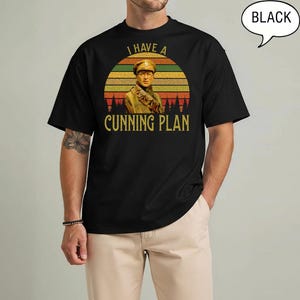 May include: Black t-shirt with a vintage-style graphic featuring a man in a military uniform, set against a sunset backdrop. The text "I HAVE A CUNNING PLAN" is printed in gold. The shirt is a classic crew neck.