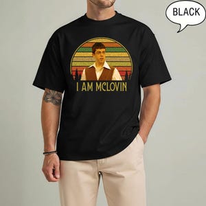 May include: Black t-shirt featuring a retro-style graphic of a man with glasses and the text "I AM MCLOVIN" in a gold font. The graphic is set against a sunset design with a circular shape. The word "BLACK" is in a speech bubble.