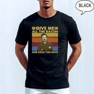 May include: Black t-shirt with a graphic featuring the text "GIVE ME ALL THE BACON AND EGGS YOU HAVE" above an image of a man. The design incorporates a retro color scheme of yellow, orange, and red stripes.