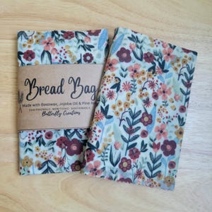 May include: Two beeswax bread bags featuring a floral pattern with red, pink, and yellow flowers on a cream background. A brown paper band wraps around one bag with the text Bread Bag, Made with Beeswax, Jojoba Oil & Pine Rosin, Butterfly Creations.