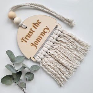 May include: A semi-circular light wood wall hanging featuring the laser-engraved text "Trust the Journey." The piece includes a cream-colored cotton cord for hanging, two wooden beads, and a textured macrame fringe attached to the bottom edge.