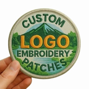 Op de afbeelding: Ronde geborduurde patch met de tekst "CUSTOM LOGO EMBROIDERY PATCHES". Het ontwerp toont een berglandschap met een rivier en bomen, in groen, blauw en oranje. De patch wordt door een hand vastgehouden.