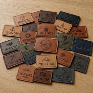 May include: A collection of rectangular leather labels in various shades of brown, tan, gray, and black. Each label features embossed icons such as bicycles, anchors, coffee cups, mountains, and tools, with the text YOUR LOGO printed on each piece.