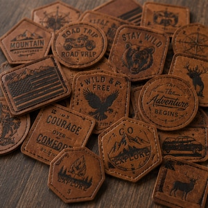 Puede incluir: Una colección de parches de cuero marrón con varios diseños en relieve, incluyendo montañas, un oso, un águila, una motocicleta, una bandera estadounidense y textos como "Stay Wild", "The Adventure Begins" y "Courage Over Comfort" en formas variadas.