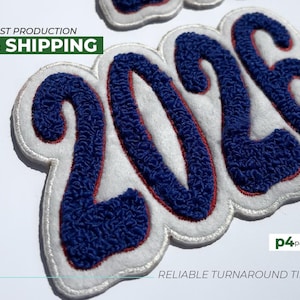 May include: Close-up of a white, blue, and red embroidered patch with the number "2026". The number is in a navy blue, textured font with a red outline, and a white border. The text "FAST PRODUCTION & SHIPPING" is in a green box. The text "RELIABLE TURNAROUND TIMES" is below.