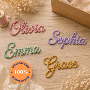 Op de afbeelding: Vier gepersonaliseerde naambordjes in cursief schrift: Olivia in roze, Sophia in paars, Emma in groen en Grace in goud. Elke naam is gemaakt van draad omwikkeld met gekleurd garen. Een oranje badge toont de tekst SATISFACTION 100% GUARANTEED.