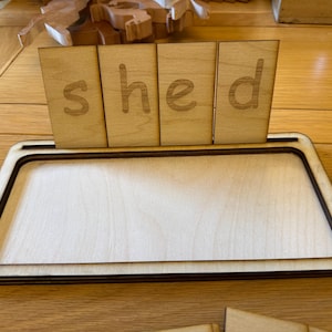 May include: A wooden educational toy displaying the word "shed" with individual letter blocks. The blocks are placed in a rectangular wooden tray. The letters are lightly etched into the wood.