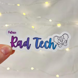 May include: A sticker with the words "future Rad Tech" in a gradient of purple and blue. A line drawing of a pelvis is on the right. The sticker is held up against a background of fairy lights.