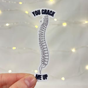May include: A white sticker with a black outline of a spine. The sticker has the words "YOU CRACK" above the spine and "ME UP" below it, all in black text. The background is a soft white with small, out-of-focus lights.