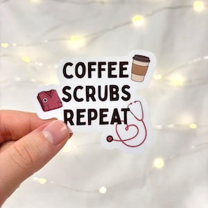 May include: A white sticker with the words "COFFEE SCRUBS REPEAT" in black text. The sticker features illustrations of a coffee cup, a medical scrub top, and a stethoscope. The sticker is held by a hand.