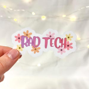 May include: A sticker with the text "RAD TECH" in pink and purple, surrounded by small flowers in shades of pink, orange, and yellow. The sticker is held against a soft, blurred background with string lights.