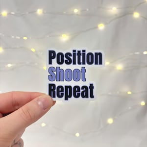 May include: A white sticker with the words "Position Shoot Repeat" in a dark blue font with a light blue shadow. The sticker is held by a hand against a background of fairy lights.