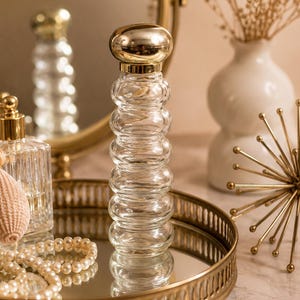 May include: A clear glass perfume bottle with a gold-colored cap and a series of rounded sections. The bottle sits on a gold-rimmed tray with other decorative items, including a mirror, a vase with dried flowers, and a pearl necklace.