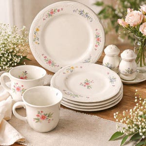 May include: A cream-colored floral dinnerware set featuring plates, cups, and salt and pepper shakers. The plates have a scalloped edge and are decorated with pink, blue, and yellow flowers. The cups and shakers have a matching floral design.