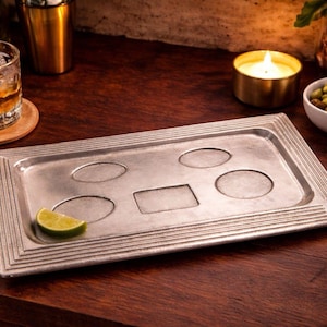 May include: A rectangular silver-colored bar tray with recessed areas for glasses and a rectangular space. A lime wedge rests on the tray. The tray is on a dark wooden surface, with a glass of whiskey and a bowl of snacks nearby.