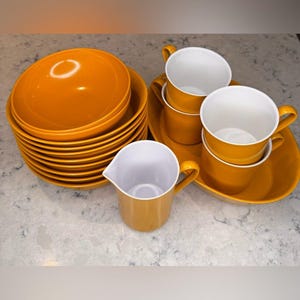 May include: A set of mustard-yellow ceramic dishes, including bowls, plates, cups, and a creamer. The bowls and plates are stacked, while the cups are arranged on a matching tray. The creamer has a handle and a spout. The cups have white interiors.