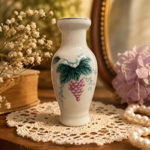May include: A small, white ceramic vase with a painted grape design. The vase features a narrow neck and a wider base. The grape design includes green leaves and a cluster of purple grapes. The vase is displayed on a lace doily.