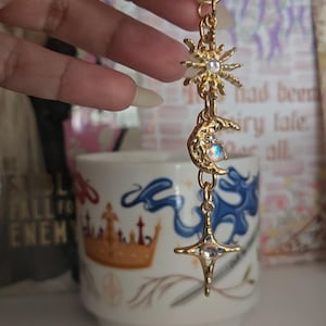 May include: Gold-colored celestial charm with a sun, crescent moon, and star design. Each charm features a small, iridescent gemstone. The charm is attached to a gold-colored clasp.