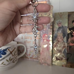May include: Silver-toned celestial charm featuring a sun, moon, and star design. Each charm has a small, round, iridescent stone at its center. The charm is suspended from a metal clasp.