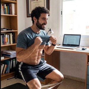 May include: A person doing squats with a resistance band around their knees. They are wearing a blue t-shirt and black shorts with a white stripe. A laptop and desk are in the background.