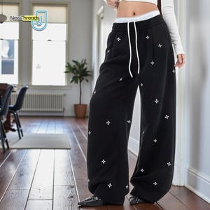 Könnte beinhalten: Schwarze weite Jogginghose mit weißer Kreuzstickerei. Die Hose hat einen Kordelzug in der Taille und einen weißen Bund. Das NewThreads-Logo ist sichtbar. Die Hose wird mit schwarzen Schuhen getragen.