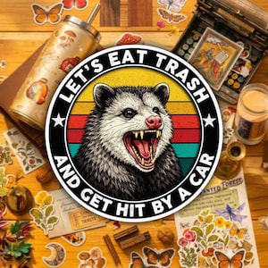 May include: A round sticker featuring an opossum with an open mouth, set against a retro sunset design. The sticker reads "LET'S EAT TRASH AND GET HIT BY A CAR" in a circular layout. The image includes a tumbler, stickers, and a vintage typewriter.